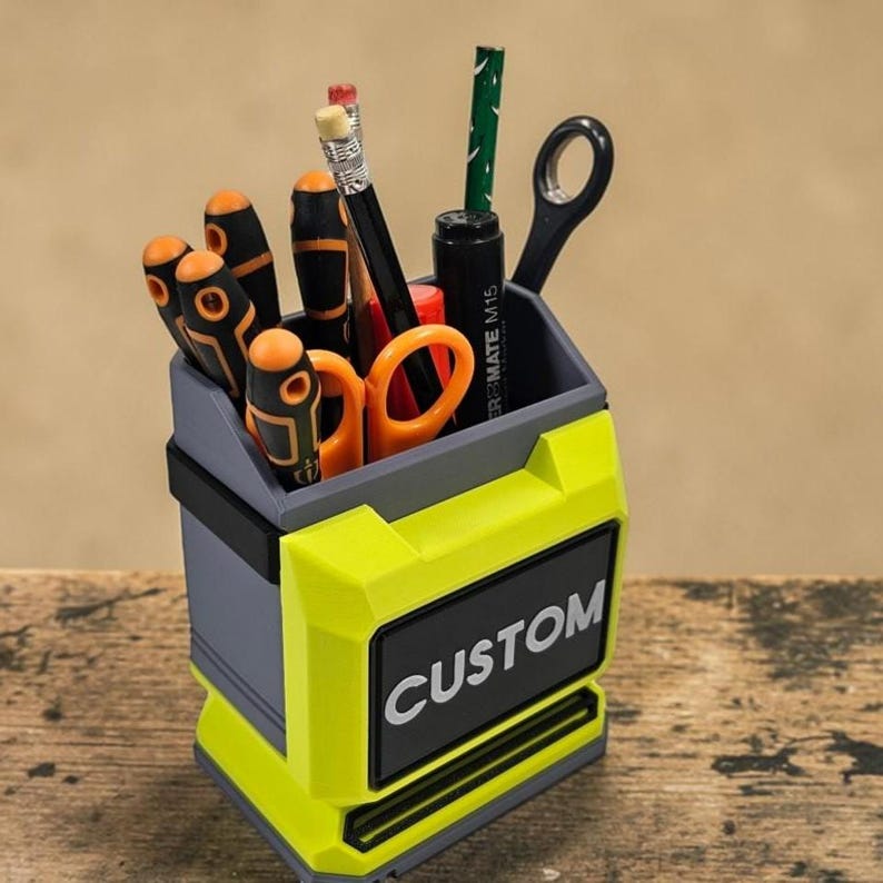 May include: A custom desk tidy in grey and neon yellow, containing tools and stationery. The organiser has the word "CUSTOM" in white on a black label. It holds screwdrivers, scissors, pencils, and a pen. The design resembles a toolbox.
