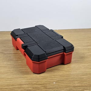 May include: A small, red and black plastic storage box with a hinged lid. The box is closed and sitting on a wooden surface.
