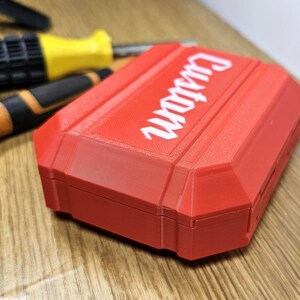 May include: A red plastic case with the word "Guardian" printed in white on the top. The case is closed and has a textured surface.