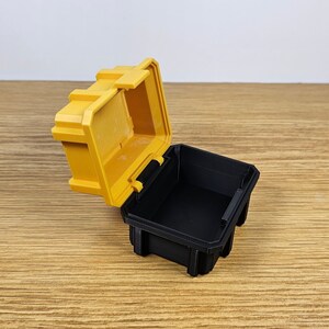 May include: A small yellow plastic storage box with a black lid. The lid is open and the box is empty.
