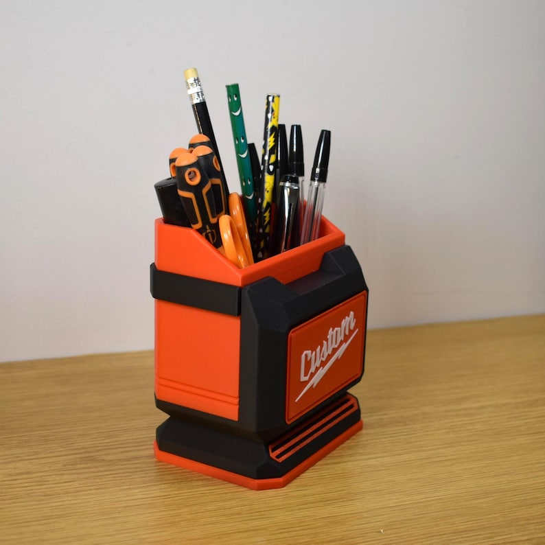 May include: An orange and black desk organizer with the word "Custom" on the side. The organizer is filled with pens, pencils, and other office supplies. The design is reminiscent of a toolbox.