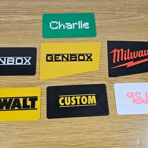 May include: Seven different name tags with various company logos and names. The tags are black, yellow, green, and white with white, black, and red lettering. The names on the tags are "Charlie", "GENBOX", "GENBOX", "DEWALT", "CUSTOM", "Milwaukee", and "ISLA".