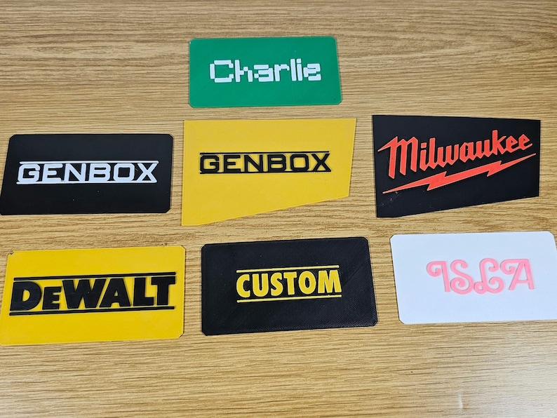 May include: Seven different name tags with various company logos and names. The tags are black, yellow, green, and white with white, black, and red lettering. The names on the tags are "Charlie", "GENBOX", "GENBOX", "DEWALT", "CUSTOM", "Milwaukee", and "ISLA".