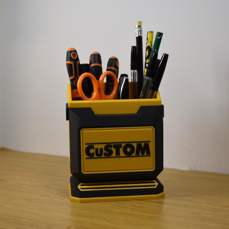 May include: A black and yellow desk organizer with the word "CUSTOM" on the front. The organizer holds various office supplies, including scissors, pens, pencils, and screwdrivers. The design is a mix of black and yellow.