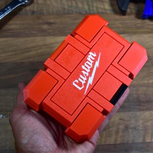 May include: Bright orange custom tool case with the word "Custom" in white script. The case is rectangular with a segmented design and a black accent. The case is held in a hand.