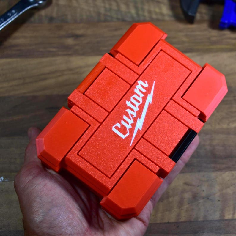 May include: Bright orange custom tool case with the word "Custom" in white script. The case is rectangular with a segmented design and a black accent. The case is held in a hand.