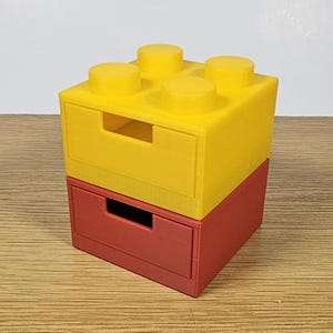 May include: Two stacked storage boxes shaped like Lego bricks. The top box is yellow with a drawer and the bottom box is red with a drawer.