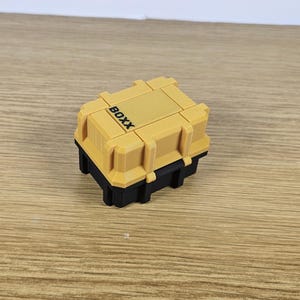 May include: A small, yellow plastic storage box with a black lid. The box has the word "BOXX" printed on the top.