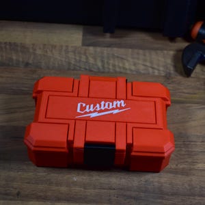 May include: A red tool case with the word "Custom" printed in white. The case has a black latch and is made of a durable material. The case is designed to hold tools and other small items.