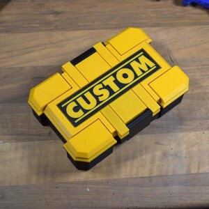 以下が含まれることがあります： 黒い背景に太字の黄色い文字で「CUSTOM」という文字が入った、黄色と黒の長方形の収納ボックス。ボックスはテクスチャ加工された表面で、プラスチック製です。