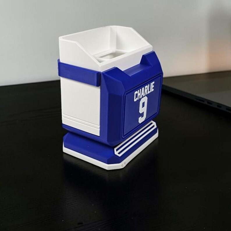 May include: A blue and white 3D-printed desk organizer with the name "Charlie" and the number "9" on the front. The organizer has a rectangular shape with a sloped top and a small opening.