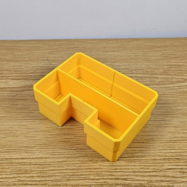 May include: A bright yellow, L-shaped desk organizer with multiple compartments. The organizer is made of a solid material and sits on a wooden surface. The design is simple and functional, suitable for organizing office supplies or small items.