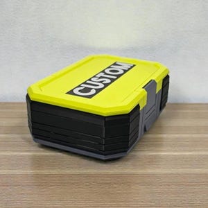 May include: A rectangular storage box with a bright yellow lid and black body. The lid features the word "CUSTOM" in bold white letters. The box has gray accents and a layered design, suggesting a durable construction.