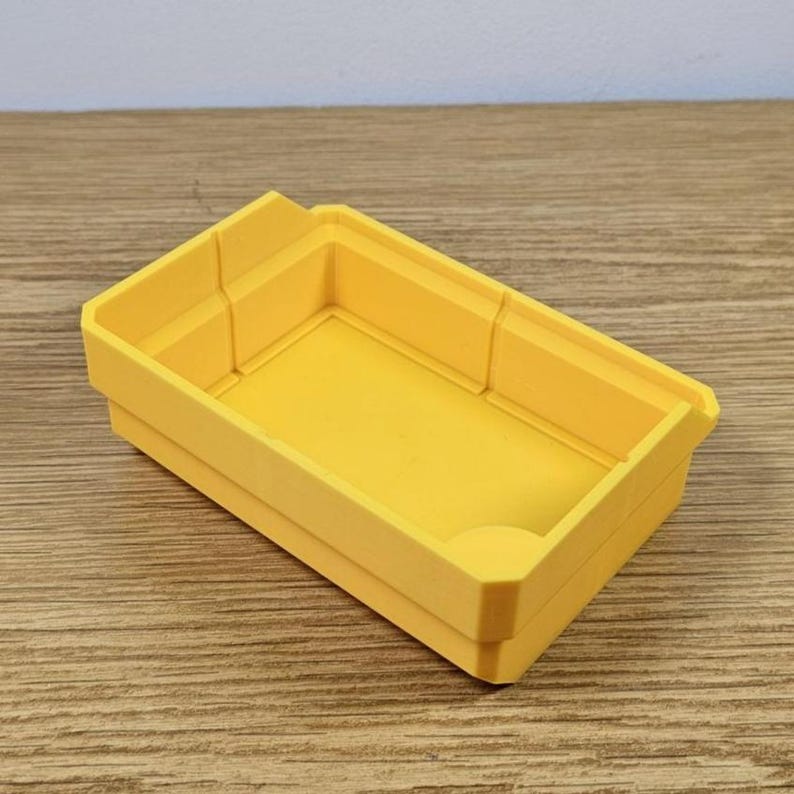 May include: A bright yellow rectangular storage bin with a slightly textured surface. The bin has a low profile with raised sides and a small notch on one corner. The bin is sitting on a wooden surface.