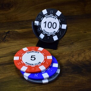Może przedstawiać: Żetony do pokera w kolorze czerwonym, białym i niebieskim, z czarnym uchwytem na żetony. Żetony wyświetlają tekst "Charlie's Poker Club" i wartości "5" i "100". Żetony leżą na drewnianej powierzchni.