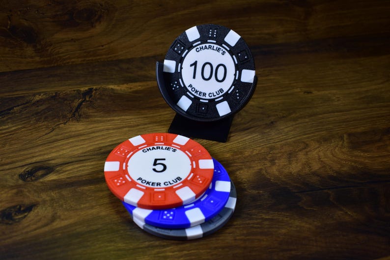Może przedstawiać: Żetony do pokera w kolorze czerwonym, białym i niebieskim, z czarnym uchwytem na żetony. Żetony wyświetlają tekst "Charlie's Poker Club" i wartości "5" i "100". Żetony leżą na drewnianej powierzchni.