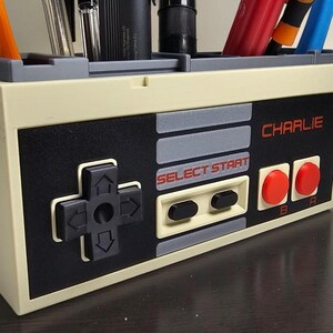 May include: A beige and black desk organizer designed to resemble a classic video game controller. It features a directional pad, buttons, and the text "SELECT START" and "CHARLIE". The organizer holds pens and pencils.