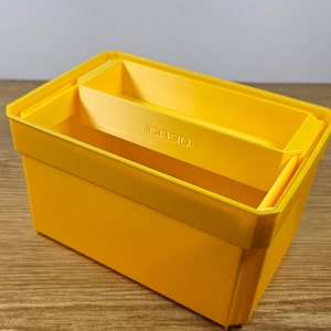 Op de afbeelding: Een felgele plastic opbergdoos met een kleinere gele doos erin. De kleinere doos is ontworpen om in de grotere doos te passen, waardoor een gelaagd opbergsysteem ontstaat.
