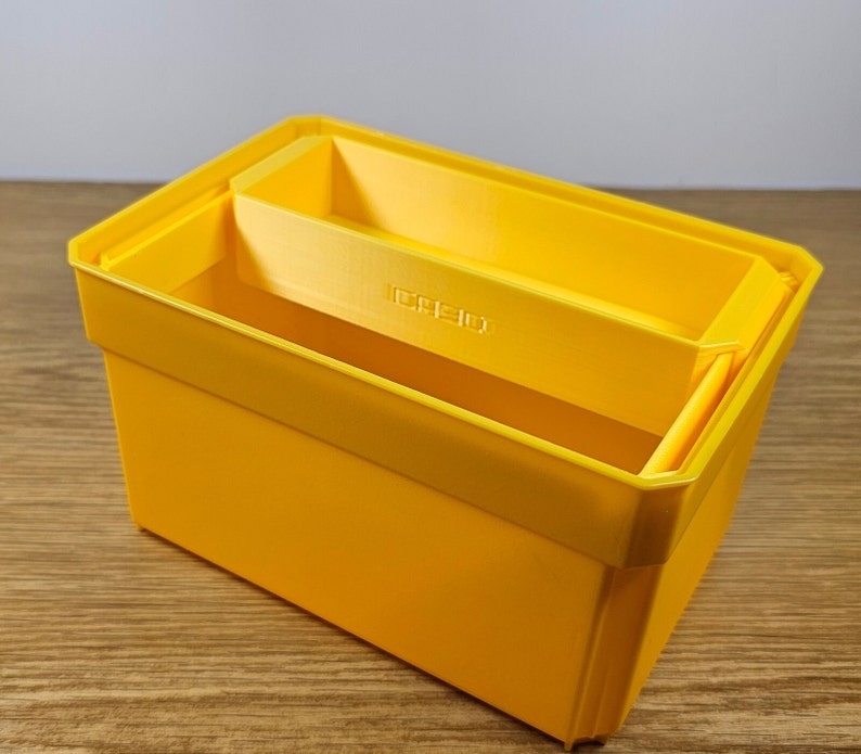 Op de afbeelding: Een felgele plastic opbergdoos met een kleinere gele doos erin. De kleinere doos is ontworpen om in de grotere doos te passen, waardoor een gelaagd opbergsysteem ontstaat.