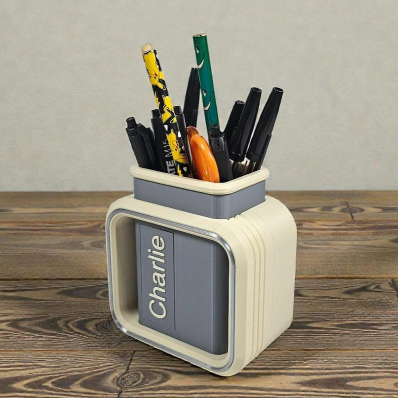 May include: A cream-colored desk organizer with a gray interior and a silver trim, holding various pens and pencils. The organizer has the name "Charlie" printed on the front.