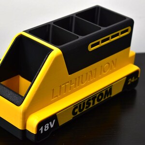 Puede incluir: Un organizador de escritorio amarillo y negro con forma de bater&iacute;a. Tiene cuatro compartimentos en la parte superior y un compartimento lateral abierto. El texto "LITHIUM ION" y "CUSTOM" son visibles, junto con "18V" y "24."