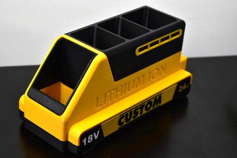 Puede incluir: Un organizador de escritorio amarillo y negro con forma de bater&iacute;a. Tiene cuatro compartimentos en la parte superior y un compartimento lateral abierto. El texto "LITHIUM ION" y "CUSTOM" son visibles, junto con "18V" y "24."