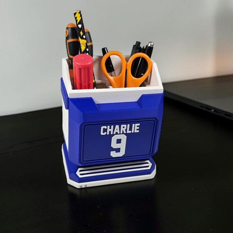 May include: A blue and white desk organizer with the name "CHARLIE" and the number "9" on the front. The organizer holds various office supplies, including scissors, pens, and screwdrivers. The organizer is made of plastic.