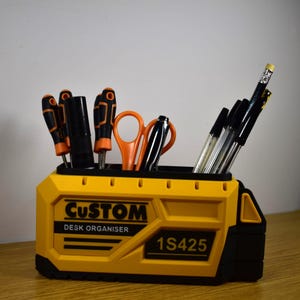K&ouml;nnte beinhalten: Ein gelb-schwarzer Schreibtisch-Organizer mit dem Text "CUSTOM DESK ORGANISER" und "1S425". Er enth&auml;lt Schraubendreher, Scheren, Stifte und einen Bleistift. Der Organizer ist f&uuml;r B&uuml;robedarf und Werkzeuge konzipiert.