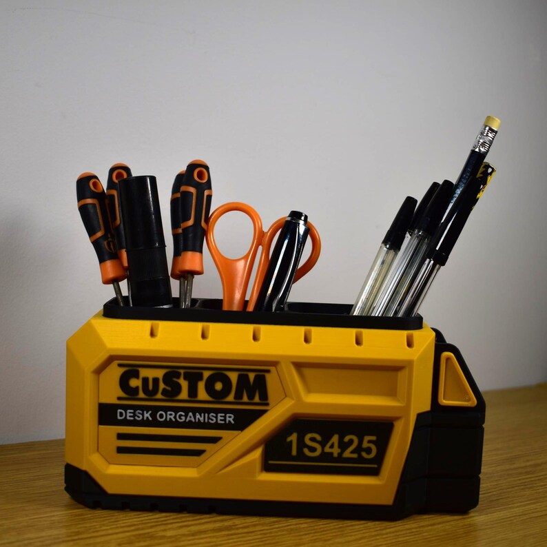 K&ouml;nnte beinhalten: Ein gelb-schwarzer Schreibtisch-Organizer mit dem Text "CUSTOM DESK ORGANISER" und "1S425". Er enth&auml;lt Schraubendreher, Scheren, Stifte und einen Bleistift. Der Organizer ist f&uuml;r B&uuml;robedarf und Werkzeuge konzipiert.