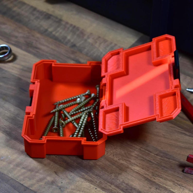 May include: An open, bright orange plastic toolbox filled with several gold-colored screws. The toolbox is rectangular with a hinged lid and sits on a wooden surface. The lid is partially open, revealing the contents.