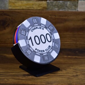 Może przedstawiać: Stos żeton&oacute;w do pokera w czarnym stojaku. G&oacute;rny żeton jest biały z czarnym napisem "Charlie's 1000 Poker Club". Żetony mają czerwone, niebieskie i białe krawędzie.
