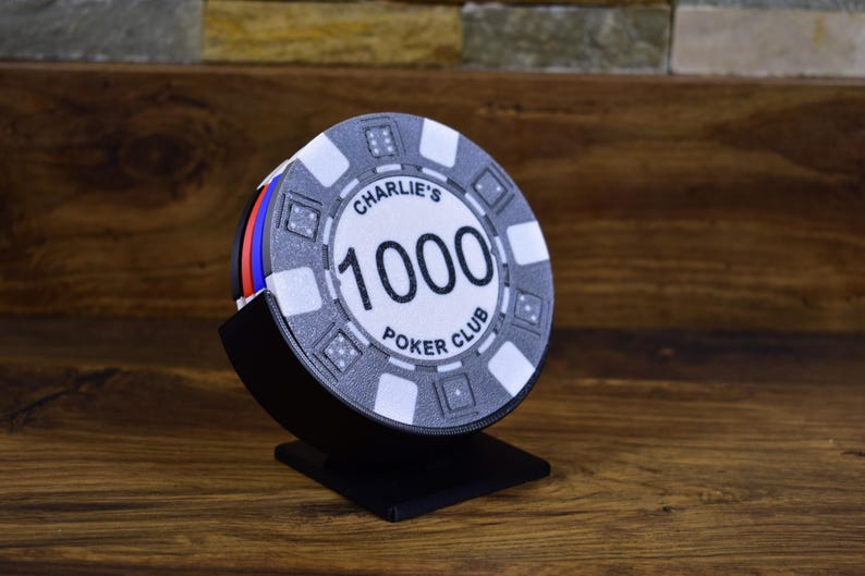 Może przedstawiać: Stos żeton&oacute;w do pokera w czarnym stojaku. G&oacute;rny żeton jest biały z czarnym napisem "Charlie's 1000 Poker Club". Żetony mają czerwone, niebieskie i białe krawędzie.