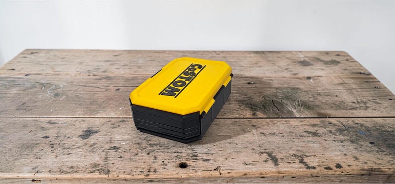 May include: A yellow and black rectangular box with the word "CUSTOM" printed on the top in black. The box is sitting on a weathered wooden surface.