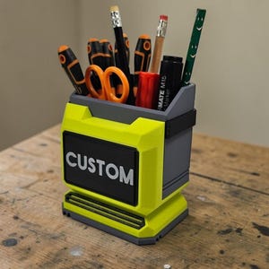 May include: A yellow and grey desk tidy with the word "CUSTOM" in white. It holds stationery items including pencils, pens, and scissors. The desk tidy is designed to keep a desk organised and tidy.