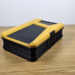 May include: A black and yellow plastic storage box with a textured lid. The box is closed and sitting on a wooden surface.