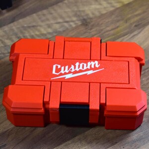 May include: A red, rectangular tool case with the word "Custom" in white script and a lightning bolt design. The case has a black handle and black latches. The case is sitting on a wooden surface.