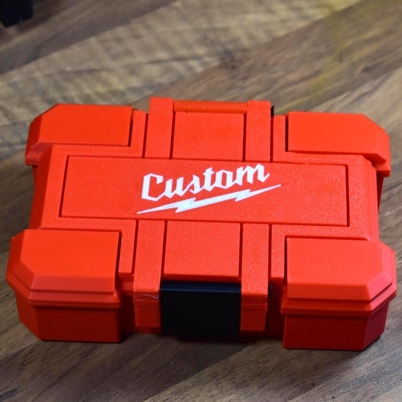 May include: A red, rectangular tool case with the word "Custom" in white script and a lightning bolt design. The case has a black handle and black latches. The case is sitting on a wooden surface.