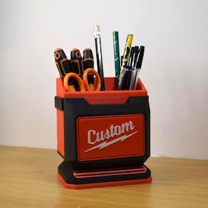 May include: A custom-made, red and black pen holder with the word "Custom" in white. The holder contains various writing utensils, including pencils, pens, and scissors. The design is reminiscent of a tool box.