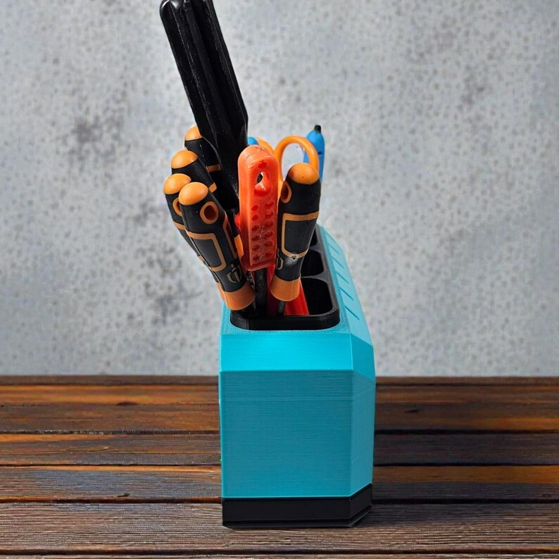 May include: A teal and black tool organizer holding various screwdrivers and tools. The organizer is made of plastic and has a geometric design. The tools have black and orange handles. The background is a gray wall and a wooden surface.