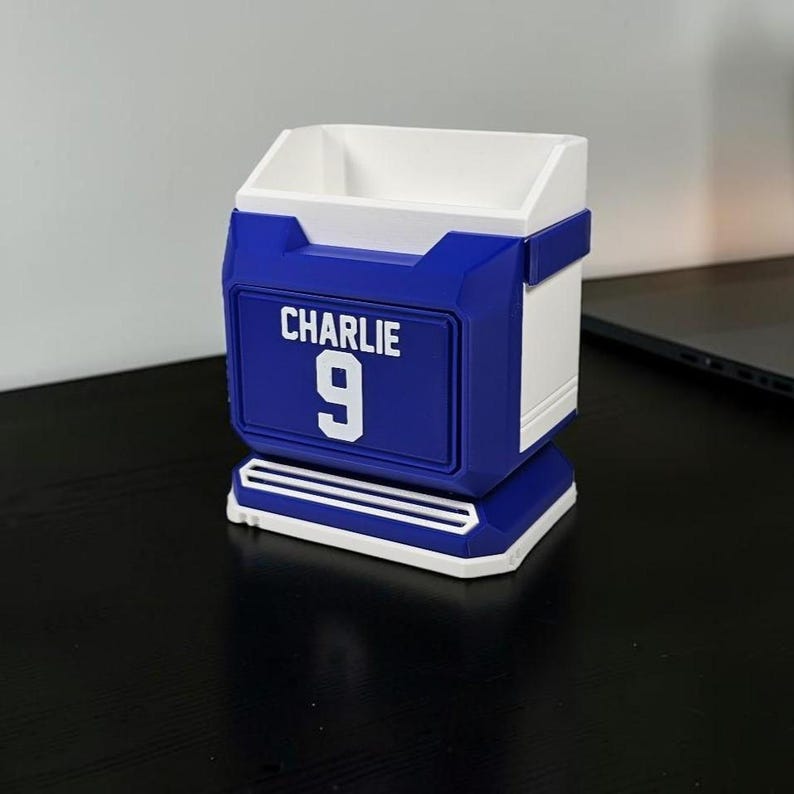 May include: A blue and white desk organizer with the name "CHARLIE" and the number "9" on the front. The organizer has a rectangular shape with a white interior and a blue exterior.