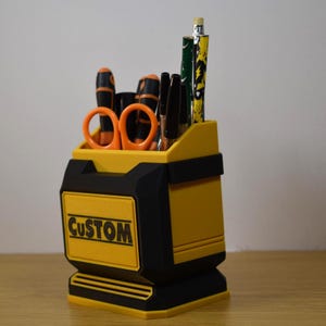 May include: A yellow and black desk organizer with the word "CUSTOM" on the front. The organizer holds orange scissors, black pens, and pencils. The design is a boxy shape with a handle-like cutout.