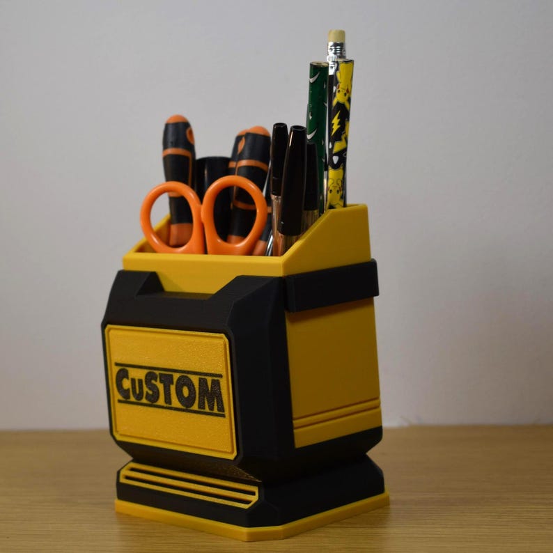 May include: A yellow and black desk organizer with the word "CUSTOM" on the front. The organizer holds orange scissors, black pens, and pencils. The design is a boxy shape with a handle-like cutout.