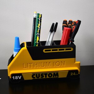 Puede incluir: Un organizador de escritorio amarillo y negro con forma de bater&iacute;a, con las palabras "LITHIUM ION" y "CUSTOM" impresas. El organizador contiene bol&iacute;grafos, l&aacute;pices y destornilladores. Una botella de pegamento azul est&aacute; en el lateral.