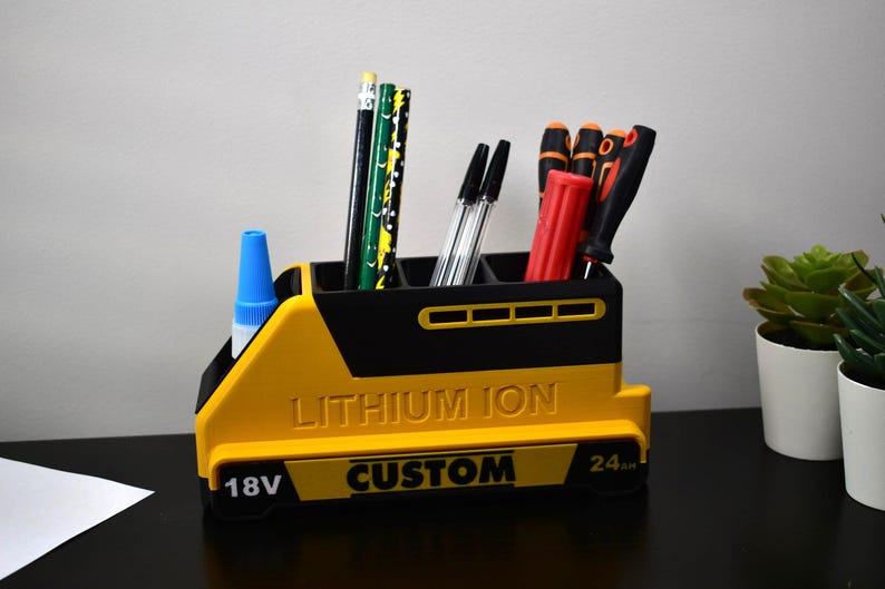 Puede incluir: Un organizador de escritorio amarillo y negro con forma de bater&iacute;a, con las palabras "LITHIUM ION" y "CUSTOM" impresas. El organizador contiene bol&iacute;grafos, l&aacute;pices y destornilladores. Una botella de pegamento azul est&aacute; en el lateral.