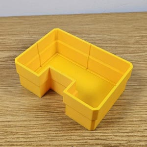 May include: A bright yellow, L-shaped plastic container. The container has a rectangular shape with raised edges and a stepped design. The object is likely used for storage or organization. The container is made of a solid, opaque material.