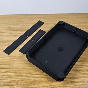 May include: A black plastic case with two black plastic inserts. The case has a rectangular shape with rounded corners and a smooth surface. The inserts are rectangular and have a textured surface.