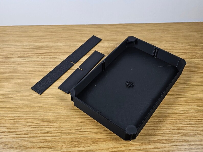 May include: A black plastic case with two black plastic inserts. The case has a rectangular shape with rounded corners and a smooth surface. The inserts are rectangular and have a textured surface.