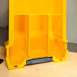 May include: A bright yellow plastic storage organizer featuring three vertical compartments separated by internal dividers. The front edge includes a black horizontal latch handle with the embossed text TOUGHSYSTEM in capital letters.