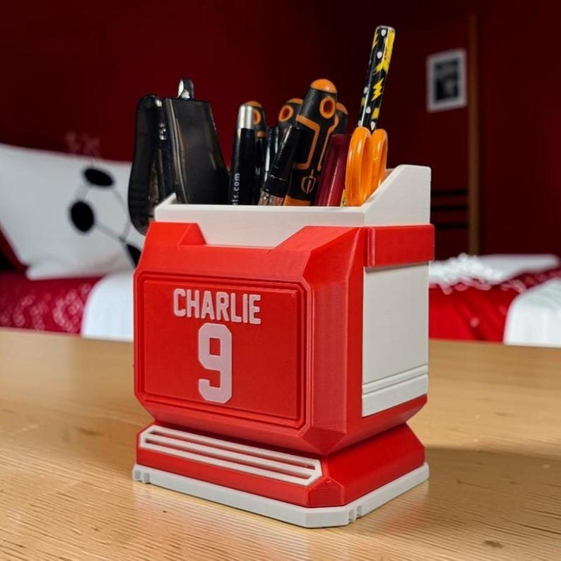 May include: A red and white desk organizer shaped like a sports jersey, featuring the name CHARLIE and the number 9 in white lettering on the front. The container holds various office supplies, including pens, a screwdriver, and orange scissors.