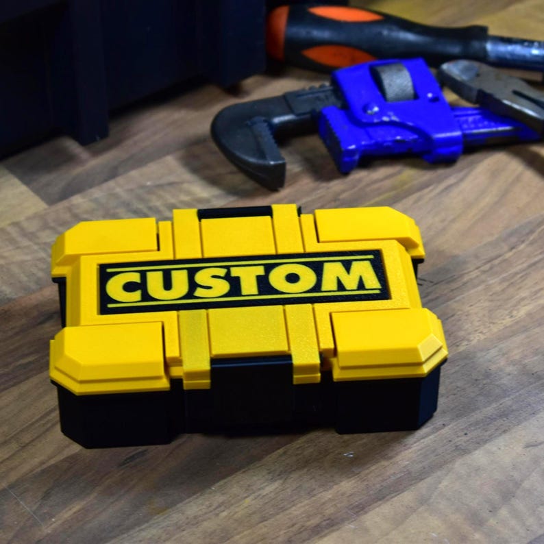 以下が含まれることがあります： 太字の黒い文字で「CUSTOM」という文字が入った、黄色と黒のツールボックス。ツールボックスは長方形で、ヒンジ付きの蓋が付いています。他のツールが背景に見えます。木製の表面にあります。